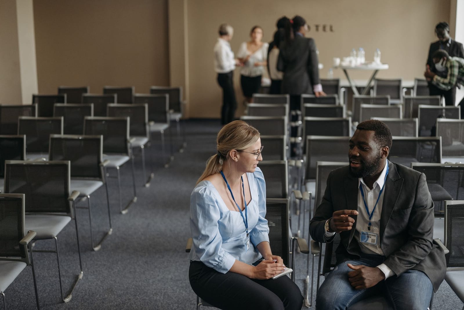 Two conference attendees wearing lanyards talk in a row of empty chairs while others gather near a refreshment table