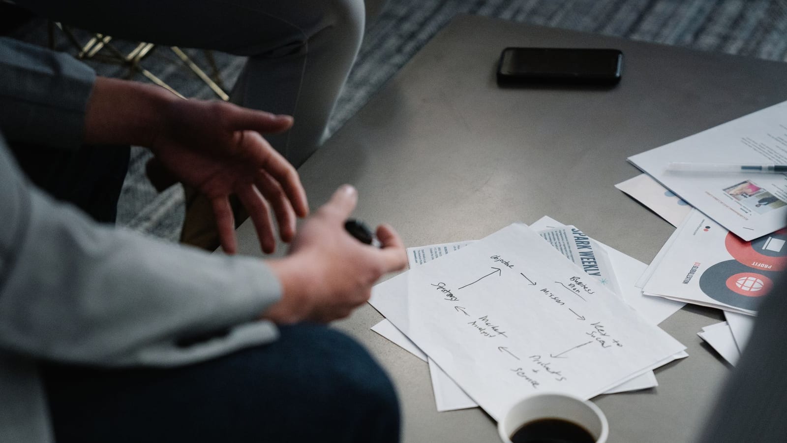 Hands gesturing over a hand-drawn strategy diagram on a meeting table, beside printed documents and a coffee cup
