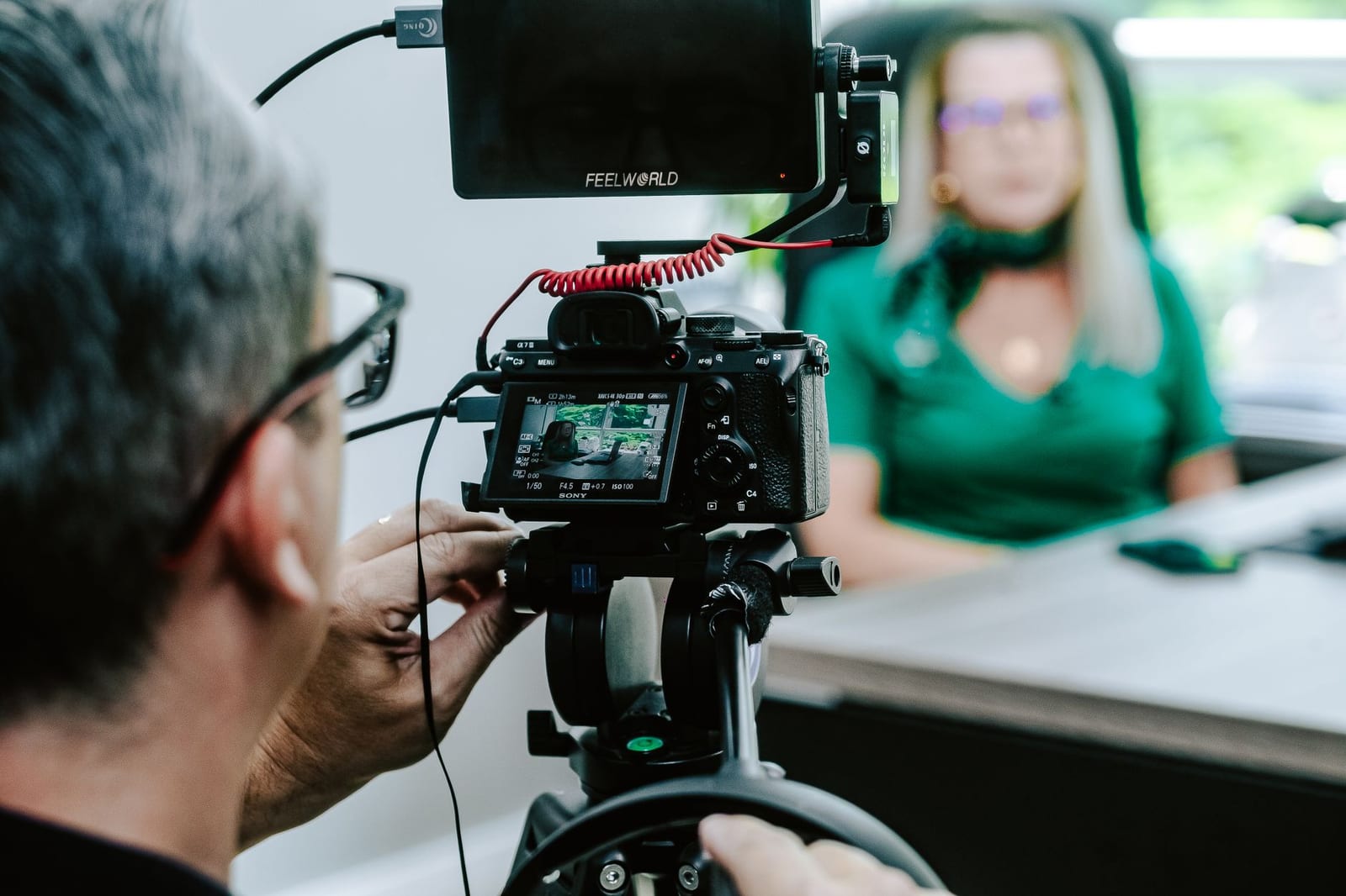 Over a camera operator&rsquo;s shoulder, a business leader sits at a desk being filmed for a video
