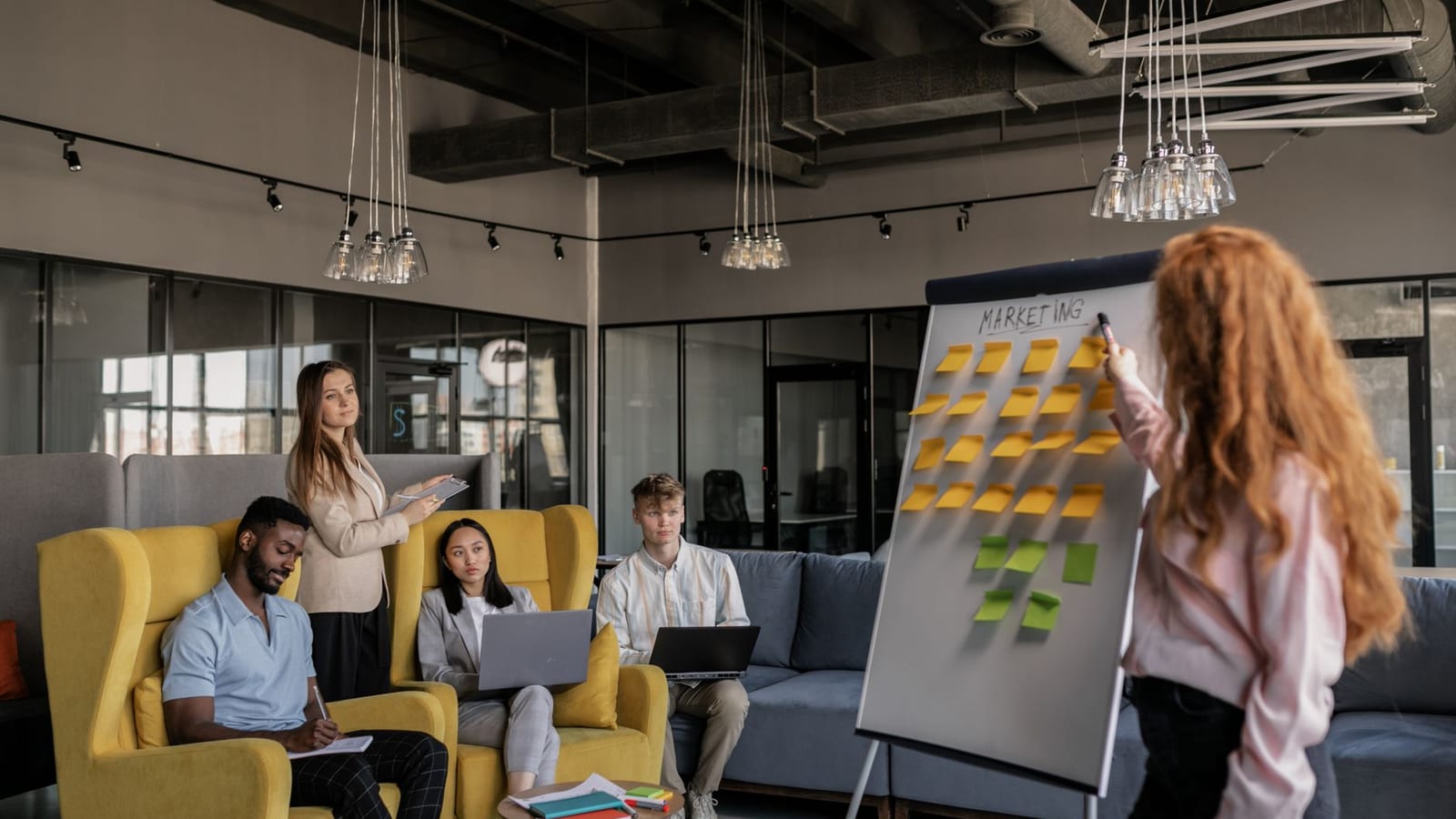 A woman points at a flip chart headed Marketing and covered in sticky notes while four colleagues watch from armchairs