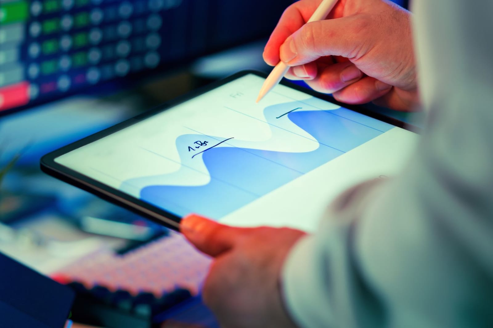 Close-up of hands marking up a blue trend chart on a tablet with a stylus, a monitor of figures behind