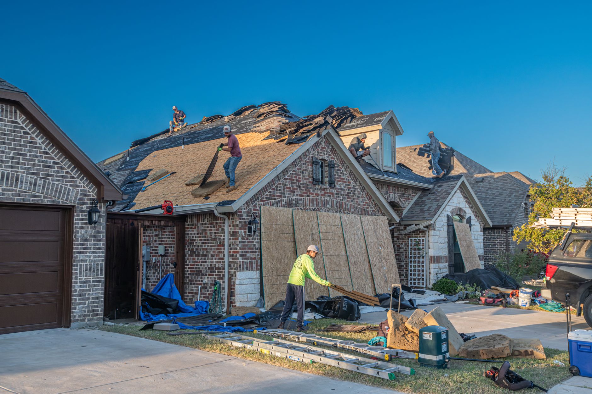 A four-person roofing crew tearing off old shingles and replacing them on a residential roof, with ladders and materials staged in the driveway below